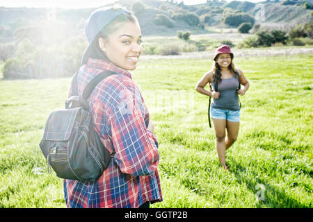 Smiling Mixed Race sisters backpacking in field near mountain Stock Photo