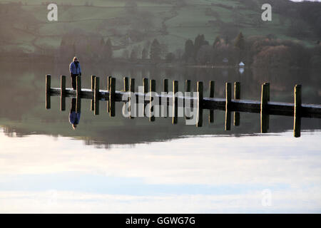 A lady wrapped up warm standing on a wooden jetty on Coniston Water on a sunny evening in winter Stock Photo
