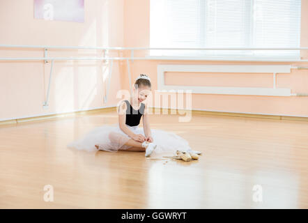 Young ballerina changing dancing shoes to pointe ones while sitting on floor Stock Photo