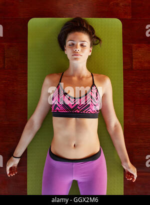 Overhead view of fit young female relaxing on yoga mat. Healthy woman in savasana yoga pose at gym. Stock Photo