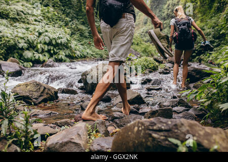 Low section shot of man and woman crossing the stream barefooted. Couple walking by the creek in forest with their shoes in hand Stock Photo