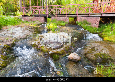 Small wooden bridge with red railings over stream with waterfall in summer park. Kotka, Finland Stock Photo