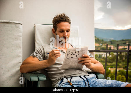 Young man sitting doing a crossword puzzle looking thoughtfully at a magazine, as he tries to think of the answer to the clue Stock Photo