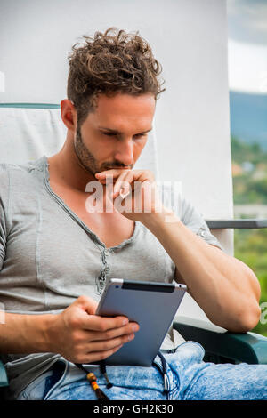 Handsome trendy man wearing t-shirt sitting and working, looking down at a tablet computer that he is holding, outdoor on house Stock Photo