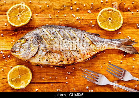Roasted gilt head bream fish on a wooden table with lemons and coarse grained salt. Stock Photo