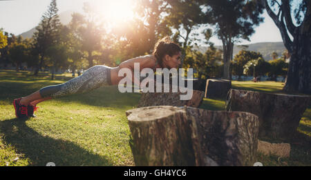 Tough young woman doing pushups on a log at park. Horizontal shot of a fit young female athlete exercising in nature. Stock Photo