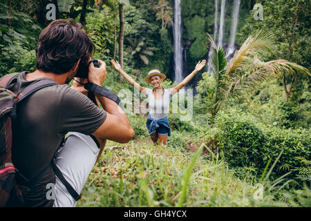 Male photographer taking pictures of a young woman standing in front of waterfall in forest. Couple enjoying a day in forest. Stock Photo