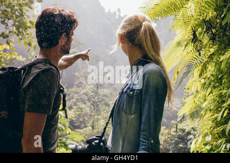 Rear view shot of young couple standing together in forest, with man pointing away. Tourist looking away in the forest. Stock Photo