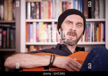 Man playing guitar at home in front of a bookshelf with books looking to camera Stock Photo