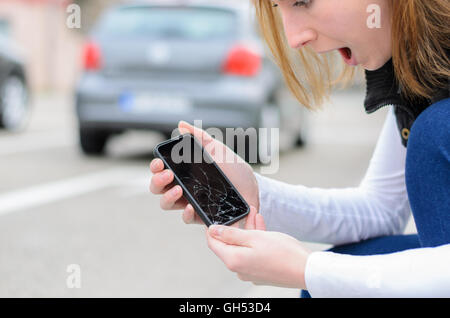 Young woman picking up her smashed mobile phone after dropping it on the cement paving of the sidewalk showing the shattered dis Stock Photo