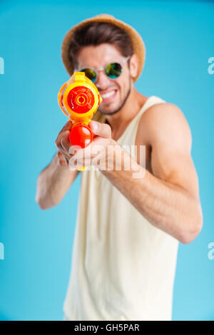 Smiling young man in hat and sunglasses playing with water gun over blue background Stock Photo