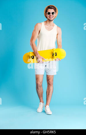 Cheerful young man in hat and sunglasses standing and holding skateboard Stock Photo