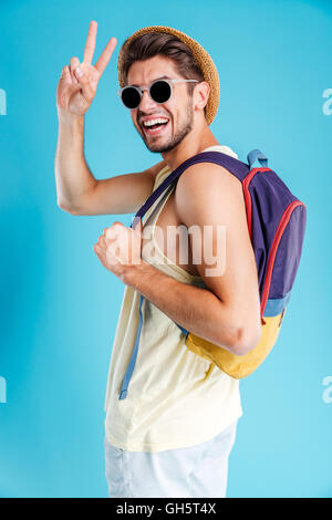 Cheerful young man in hat and sunglasses holding backpack and showing peace sign Stock Photo