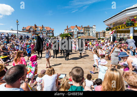 Popular 'Hobby Horse Club' event during the Broadstairs Folk Week. Ossies, iconic black bird figures on crowded deck dancing the hokey-cokey with families and children rushing on to join in. Outdoors, sunshine. Stock Photo