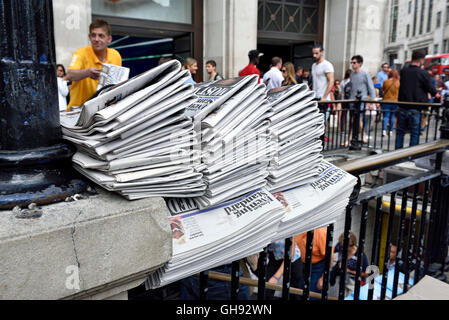 Free newspapers.  Copies of The Evening Standard piled up on railing at Oxford Circus Station, vendor in background, Central Lon Stock Photo