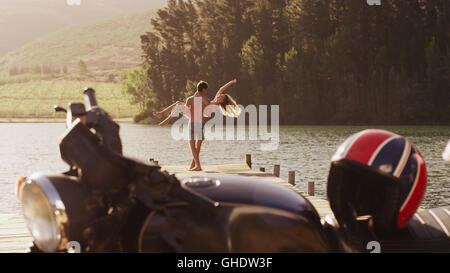 Young man carrying exuberant woman on lakeside dock behind motorcycle Stock Photo