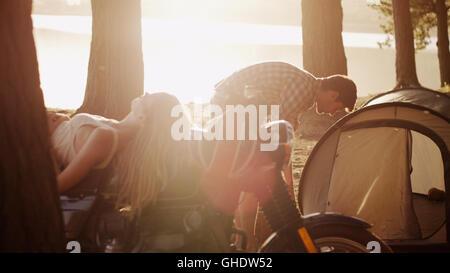 Young man preparing tent at campsite next to woman laying on motorcycle Stock Photo