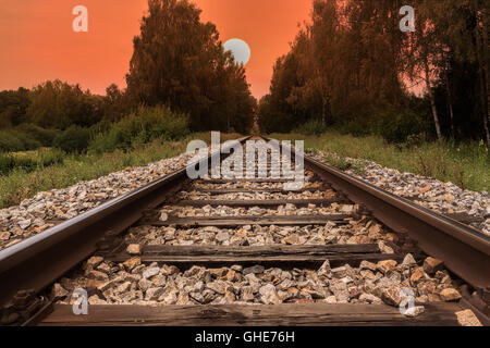 Railway line, trail or track  going through landscape at sunset or dawn. Red sky with big evening sun. Stock Photo