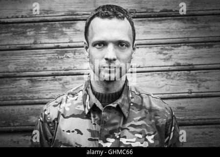 Young Caucasian military man in camouflage uniform. Closeup black and white frontal portrait over wooden wall Stock Photo