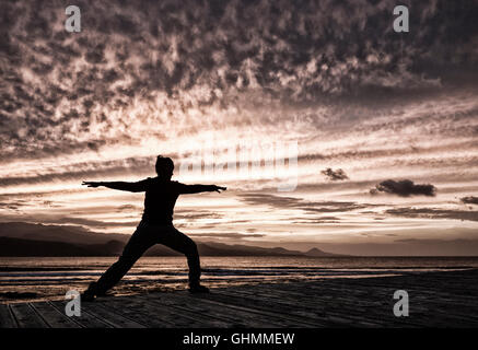 Woman practicing Yoga overlooking the sea under a dramatic sky at sunset Stock Photo