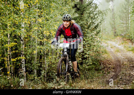 young blond woman cyclist rides through forest during Regional competitions on cross-country bike Stock Photo