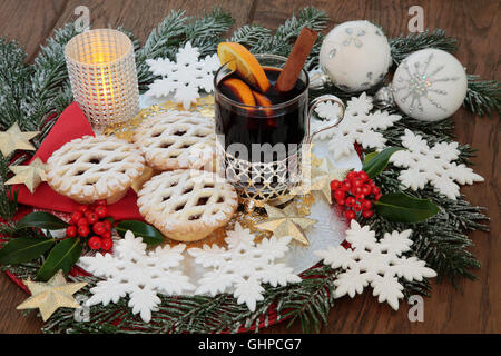 Christmas party food and drink with mince pies, mulled wine, white snowflake and gold star bauble decorations, holly and fir. Stock Photo