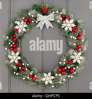 Christmas wreath decoration with silver bow, red bauble and white snowflake decorations, holly and snow covered blue spruce fir. Stock Photo