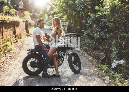 Outdoor shot of cheerful young couple with motorcycle on country road. Man sitting on motorbike with woman holding a smartphone. Stock Photo