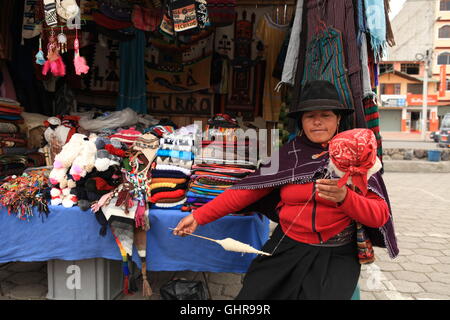 A traditionally dressed woman sits outside her handwoven fabric stall in Otavalo Market and spins cotton into thread Stock Photo