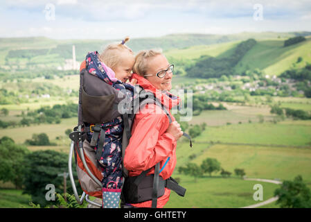 Young happy mother hold little traveler on back - baby girl in carrying backpack enjoying travel adventure, Hiking activity with Stock Photo