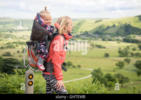 Young happy mother hold little traveler on back - baby girl in carrying backpack enjoying travel adventure, Hiking activity with Stock Photo