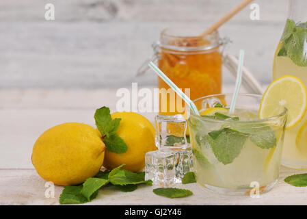 Glass of lemonade with lemon slice, mint leaves and ice cubes near lemons, lemonade bottle and honey jar on rustic white wood Stock Photo