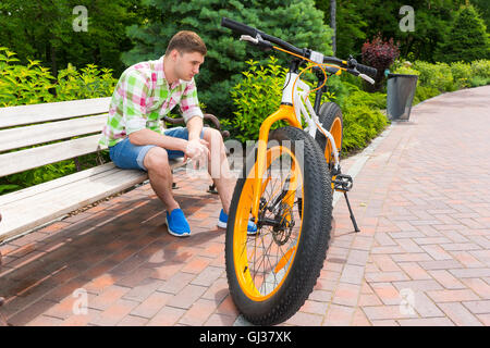 Lonely single man with sad expression sitting on bench near bike with fat tires parked on brick sidewalk in beautiful green park Stock Photo