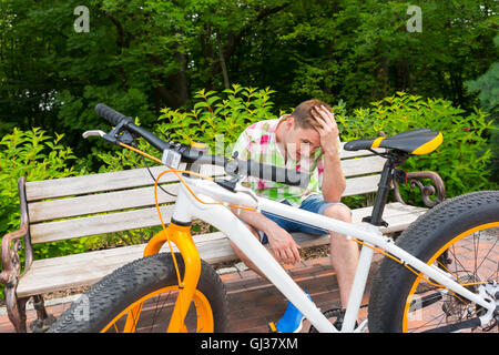 Young man with sad expression sitting on bench near bike with fat tires parked on brick sidewalk in beautiful green park Stock Photo