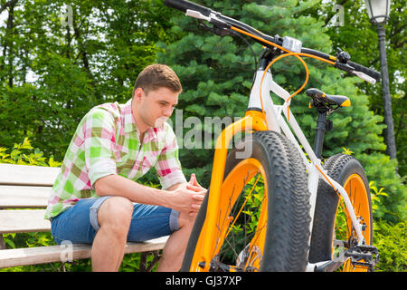 Pensive young man sitting on bench near bike with fat tires in beautiful green park Stock Photo