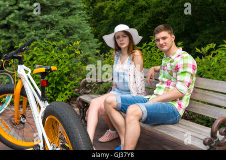 Young fashionable couple sitting on bench near bikes parked on brick sidewalk in beautiful green park Stock Photo