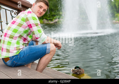 Young man feeding ducks in a pond sitting on the wooden deck in a park with fountain in a background Stock Photo