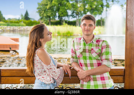 Young romantic couple leaning on railing of the wooden gazebo standing in front of human-made waterfall in a park with pond, fountain and different trees. Stock Photo