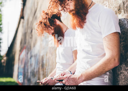 Young male hipster twins with red beards leaning against wall texting on smartphones Stock Photo