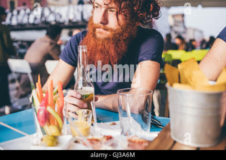 Young male hipster with red hair and beard drinking bottled beer at sidewalk bar Stock Photo