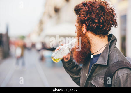 Young male hipster with red hair and beard drinking bottled beer on city street Stock Photo