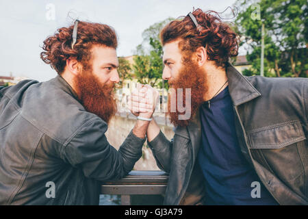 Young male hipster twins with red hair and beards arm wrestling on bridge Stock Photo