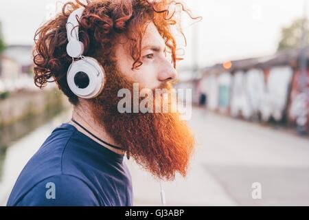 Young male hipster with red hair and beard listening to headphones in city Stock Photo