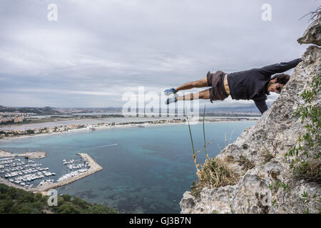 Male rock climber balancing horizontally mid air on coastal rock, Cagliari, Italy Stock Photo