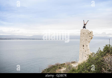 Male rock climber doing handstand on top of ruined tower on coast, Cagliari, Italy Stock Photo