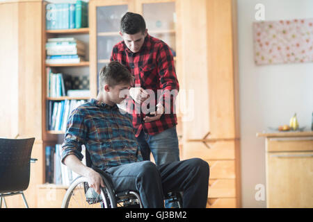 Young man using wheelchair reading smartphone texts with friend in kitchen Stock Photo