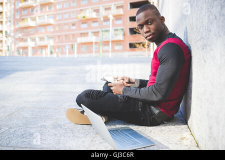 Young man sitting outdoors, leaning against wall, using digital tablet, laptop beside him Stock Photo