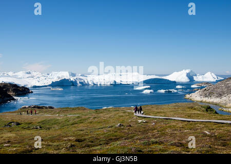 People on a boardwalk across the Sermermiut Inuit settlement archaeological site with icebergs in Disko Bay in summer. Ilulissat Greenland Stock Photo