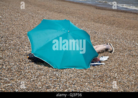 Legs protruding from beneath blue umbrella on Dunwich beach, Suffolk. Stock Photo