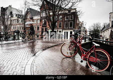 A cyclist and red bicycles with an umbrella on a bridge in the rain in Amsterdam Stock Photo